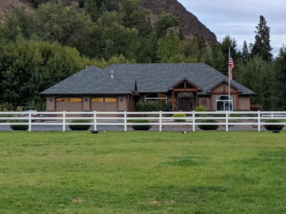 Large ranch home with a stone and timber entry, a white rail fence and a wide lawn below a forested hill