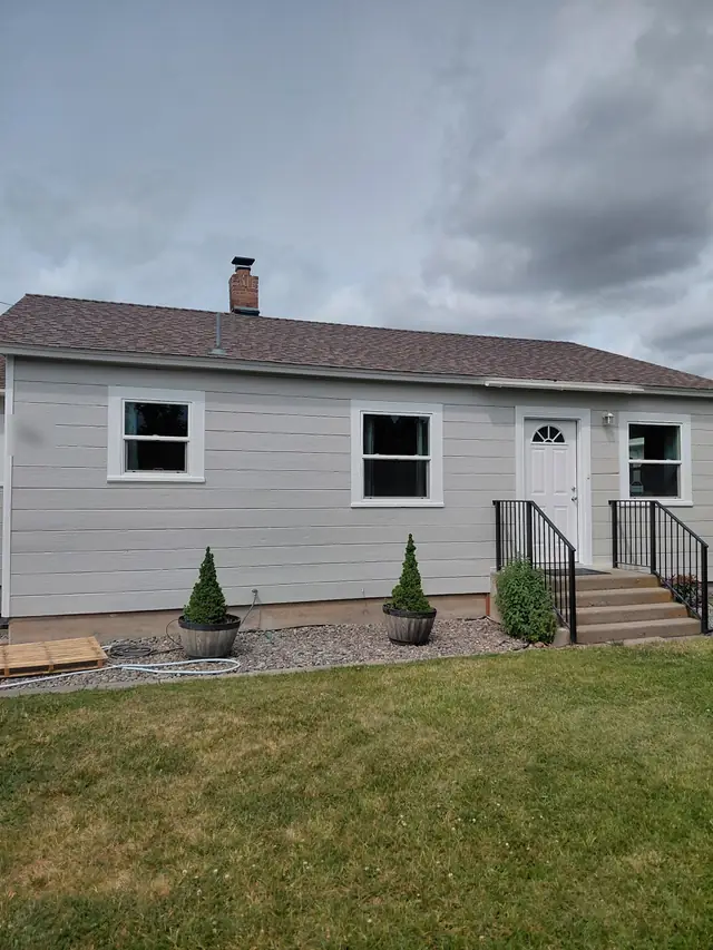 Small grey bungalow with bright white trim and windows, black iron stair rails and potted topiaries under an overcast sky